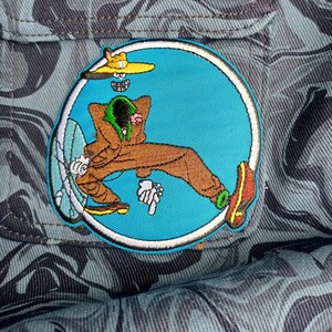 May include: Embroidered patch featuring a cartoon character in a brown jumpsuit, yellow hat, and red shoes, relaxing inside a blue circle. The patch is set against a camouflage fabric background, with a light blue and gray pattern.