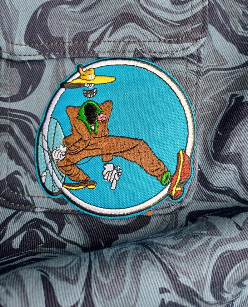 May include: Embroidered patch featuring a cartoon character in a brown jumpsuit, yellow hat, and red shoes, relaxing inside a blue circle. The patch is set against a camouflage fabric background, with a light blue and gray pattern.