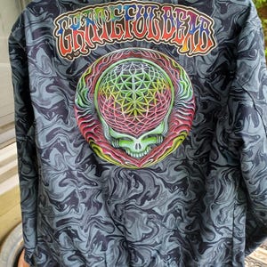 May include: A dark blue jacket with a swirling pattern. The back features a colorful graphic of a skull and geometric design, with the text "Grateful Dead" above it. The jacket has a drawstring at the bottom.