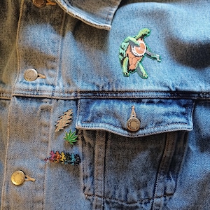 Könnte beinhalten: Hellblaue Jeansjacke mit mehreren Anstecknadeln. Die Anstecknadeln zeigen eine grüne Schildkröte, die Banjo spielt, einen goldenen Blitz, ein grünes Marihuanablatt und eine regenbogenfarbene Anstecknadel mit tanzenden Bären.