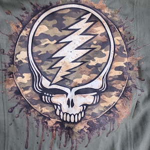 May include: Olive green jacket with a large circular patch featuring a skull and lightning bolt design. The patch has a camouflage pattern and is surrounded by brown paint splatter details.