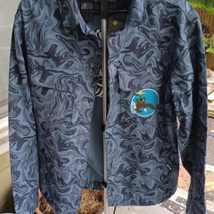 May include: A button-up shirt with a swirling blue and gray pattern. The shirt has two chest pockets and a patch with a cartoon image. The shirt is hanging on a clothing rack.
