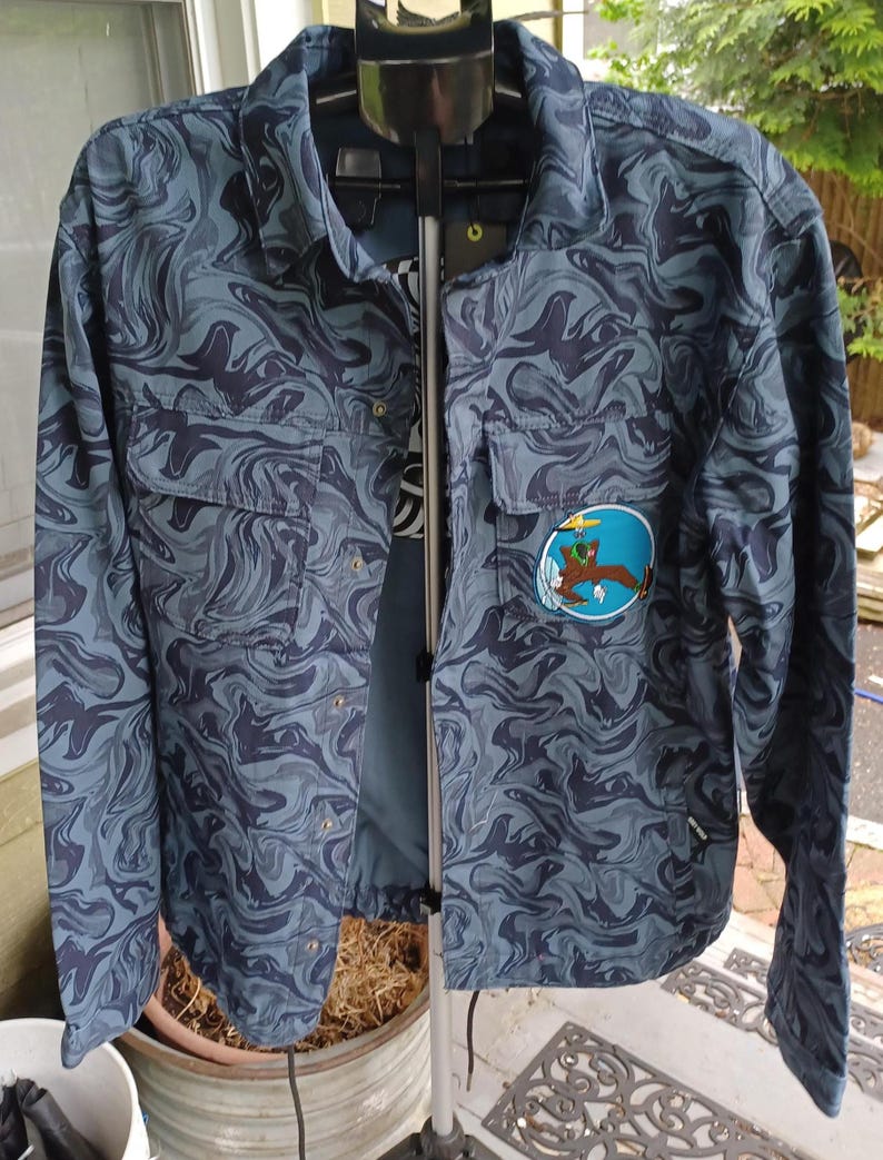 May include: A button-up shirt with a swirling blue and gray pattern. The shirt has two chest pockets and a patch with a cartoon image. The shirt is hanging on a clothing rack.