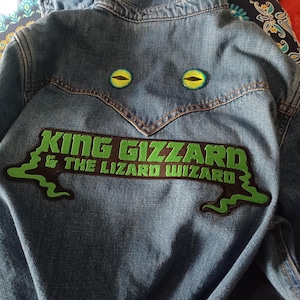 May include: A blue denim jacket with a patch displaying the words "KING GIZZARD & THE LIZARD WIZARD" in green and black. Two green and yellow eye-shaped pins are above the text.
