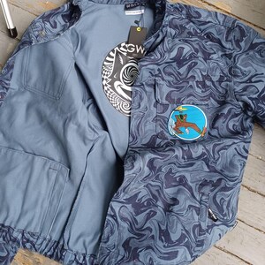 May include: A blue jacket with a swirling pattern, open to reveal a gray t-shirt with a black and white graphic. The jacket has a patch with a cartoon figure. The jacket has two front pockets and a tag that says "GW".
