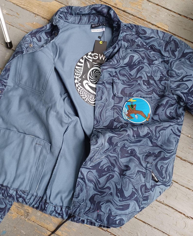 May include: A blue jacket with a swirling pattern, open to reveal a gray t-shirt with a black and white graphic. The jacket has a patch with a cartoon figure. The jacket has two front pockets and a tag that says "GW".