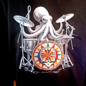 May include: Black sweatshirt featuring a white octopus playing a drum set. The drum set has silver cymbals and drums. The bass drum has a colorful design with a skull and starburst pattern.