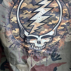 May include: Camouflage jacket featuring a Steal Your Face skull and lightning bolt design. The graphic is in a circular frame with a camouflage pattern and a white outline. The jacket has a green, brown, and tan camouflage pattern.