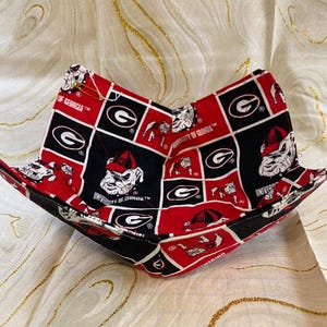 May include: A red, black, and white bowl cozy featuring the University of Georgia logo and bulldog mascot. The fabric is patterned with the university's logo and bulldog mascot. The cozy is designed to hold a bowl of food and keep it warm.
