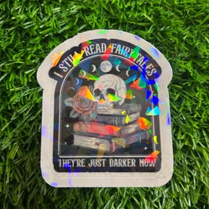 Peut inclure: Autocollant holographique avec le texte "I STILL READ FAIRYTALES THEY'RE JUST DARKER NOW". L'autocollant représente un crâne, des livres, une rose et des croissants de lune. L'autocollant a une bordure blanche et est sur une surface verte.