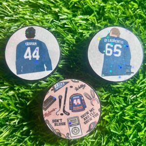 May include: Three circular hockey pucks with holographic accents. Two pucks feature a hockey player in a blue jersey with names and numbers 44 Graham and 65 Di Laurentis. The third puck displays a collage of icons including a jersey, microphone, and music player.