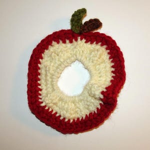 May include: A circular crochet apple slice coaster featuring a cream-colored center, a thick red outer border, and a small green leaf with a brown stem attached to the top edge. The yarn texture is visible throughout the handmade piece.