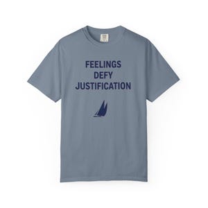 May include: A slate blue short-sleeve t-shirt featuring the dark navy text "FEELINGS DEFY JUSTIFICATION" printed in a bold, sans-serif font on the chest, positioned above a small, minimalist graphic of a sailboat with two sails.