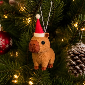 Puede incluir: Una figura de capibara marrón que lleva un pequeño gorro de Papá Noel rojo y blanco. El capibara tiene una superficie texturizada, ojos oscuros y un hocico redondeado. Está suspendido por un fino cordón plateado para colgar en el árbol.