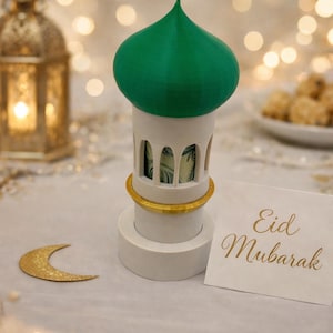 Könnte beinhalten: Ein weiß-grüner dekorativer Geldhalter in Form einer Moschee mit einem goldenen Band. Der Halter hat gewölbte Fenster und eine grüne Kuppel. Eine Karte mit "Eid Mubarak" liegt daneben.
