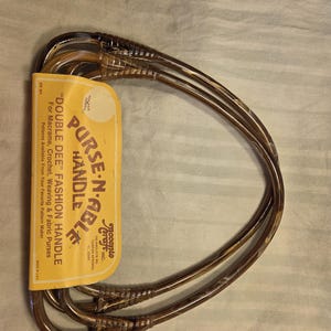 May include: Brown "Double Dee" purse handles with a yellow label. The label reads "PURSE-N-ABLE" and "DOUBLE DEE FASHION HANDLE" with text about macrame, crochet, weaving, and fabric purses. The handles are made of a clear, brown plastic.