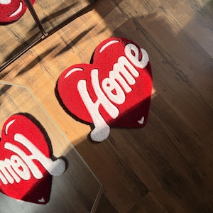 May include: Red heart-shaped rug with the word "Home" in white script. The rug is made of a soft, textured material and is placed on a wooden floor. The rug is approximately 24 inches wide and 24 inches long.