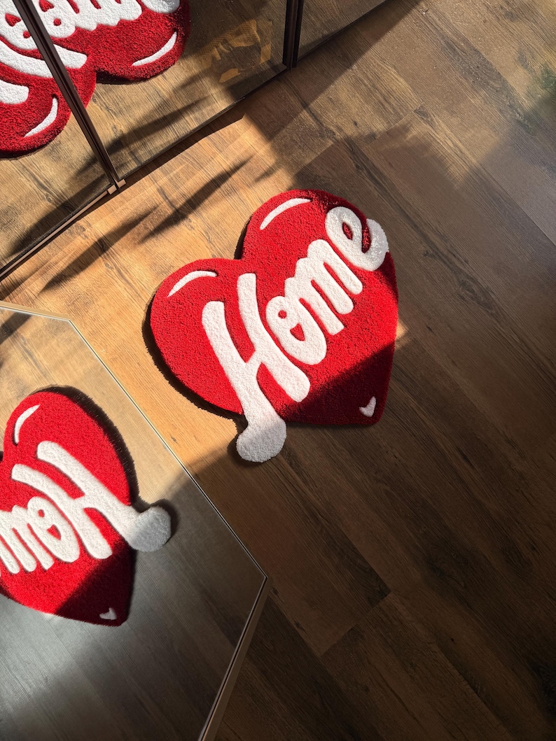 May include: Red heart-shaped rug with the word "Home" in white script. The rug is made of a soft, textured material and is placed on a wooden floor. The rug is approximately 24 inches wide and 24 inches long.