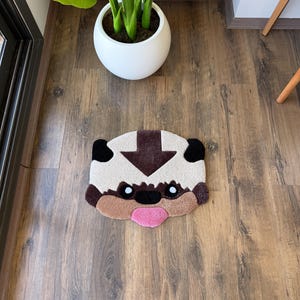 May include: A beige and brown rug shaped like a cartoon character's face, with black ears, a brown arrow, and a pink tongue. The rug is on a wooden floor, near a white potted plant.