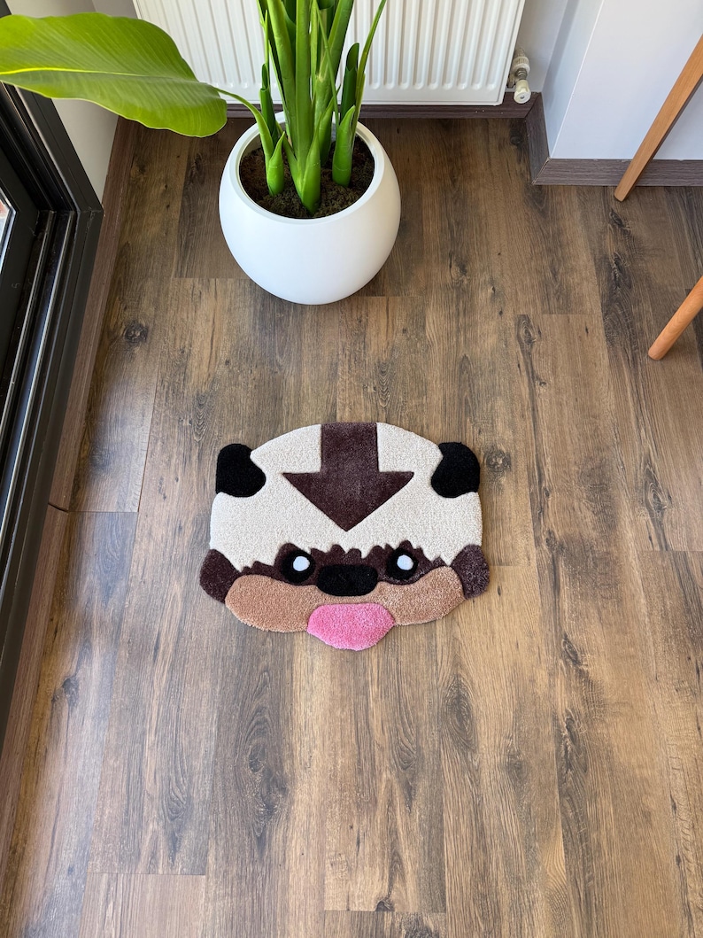 May include: A beige and brown rug shaped like a cartoon character's face, with black ears, a brown arrow, and a pink tongue. The rug is on a wooden floor, near a white potted plant.