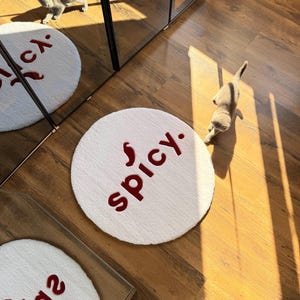 May include: A white, round rug with the word "spicy" in red lettering. The rug is on a wooden floor, and a gray kitten is walking nearby. The rug is a decorative item.