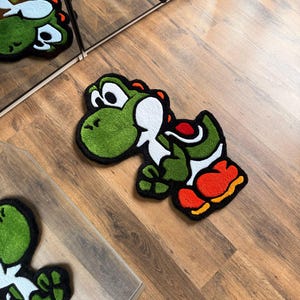 May include: A green, white, red, and orange Yoshi rug. The rug is in the shape of the character Yoshi from the Super Mario video game series. The rug is on a wooden floor.