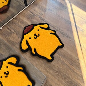 May include: Yellow rug in the shape of a cartoon dog with a brown hat and black outline. The rug has a smiling face and is placed on a wooden floor. A second rug is partially visible.