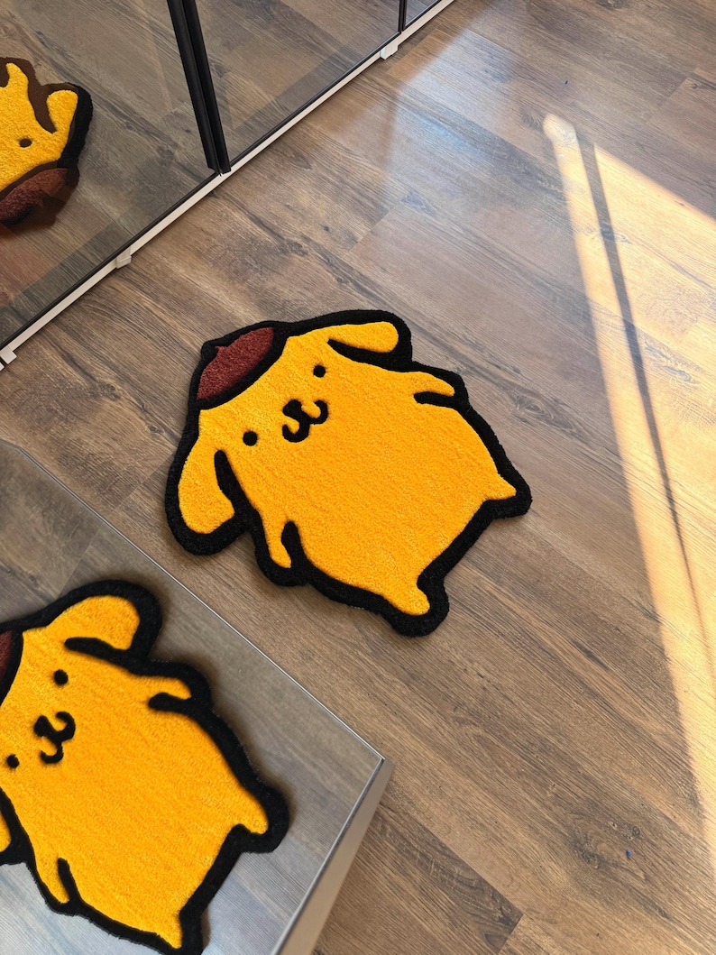 May include: Yellow rug in the shape of a cartoon dog with a brown hat and black outline. The rug has a smiling face and is placed on a wooden floor. A second rug is partially visible.