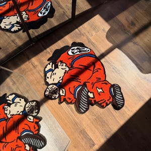 May include: A red and orange rug featuring a cartoon character in a kneeling position. The rug has black, white, and blue accents, and is placed on a wooden floor. The image is taken from an elevated angle, with a mirror reflecting the rug.