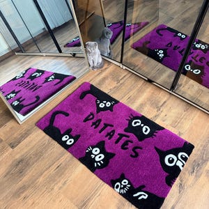 May include: A purple doormat with black cat illustrations and the word "PATATES" in white. The doormat is rectangular and placed on a wooden floor. The image also shows a cat and a mirror.