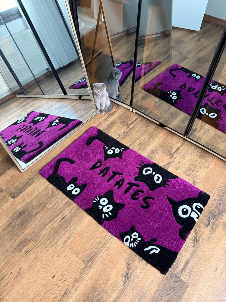 May include: A purple doormat with black cat illustrations and the word "PATATES" in white. The doormat is rectangular and placed on a wooden floor. The image also shows a cat and a mirror.