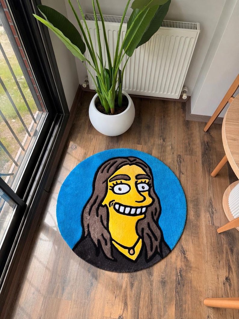 May include: A round, blue rug featuring a cartoon character with yellow skin, brown hair, and a black outfit. The rug is placed on a wooden floor, with a white potted plant and a window in the background.