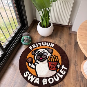 May include: Round brown rug featuring a cartoon pug holding fries and a meatball, with the text "FRITUUR SWA BOULET" in white. The pug is set against an orange circle, with a brown border. The rug is on a wooden floor.
