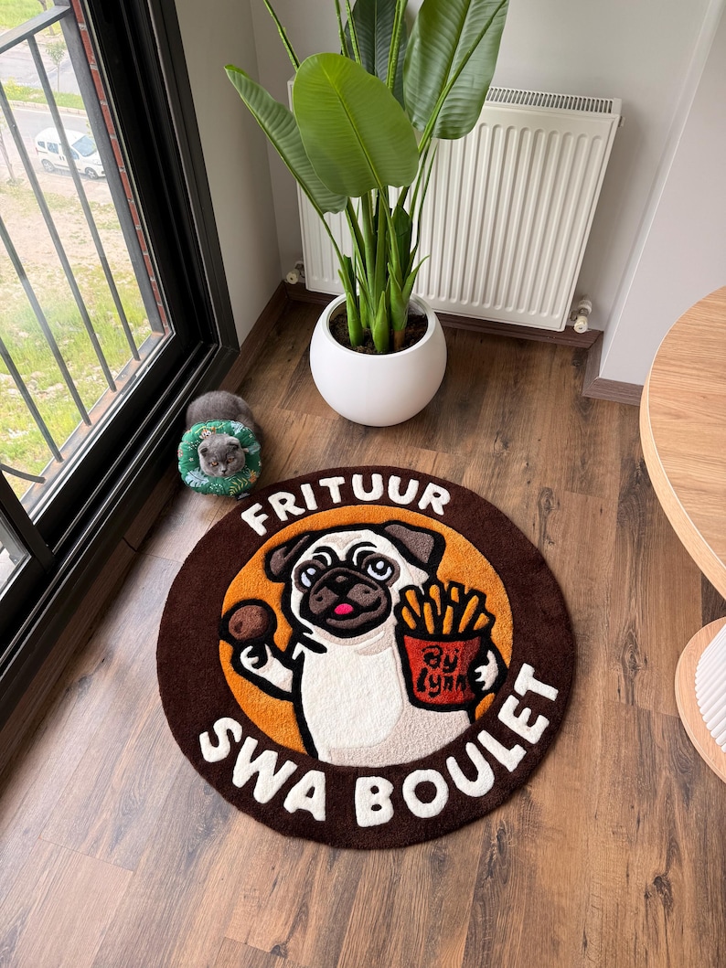 May include: Round brown rug featuring a cartoon pug holding fries and a meatball, with the text "FRITUUR SWA BOULET" in white. The pug is set against an orange circle, with a brown border. The rug is on a wooden floor.