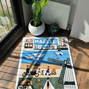May include: A rectangular rug with a whimsical design featuring a town scene with a windmill, airplane, and people. The rug has a white border and the name "MAELLE" is displayed at the top. The color palette includes blue, green, beige, and brown.