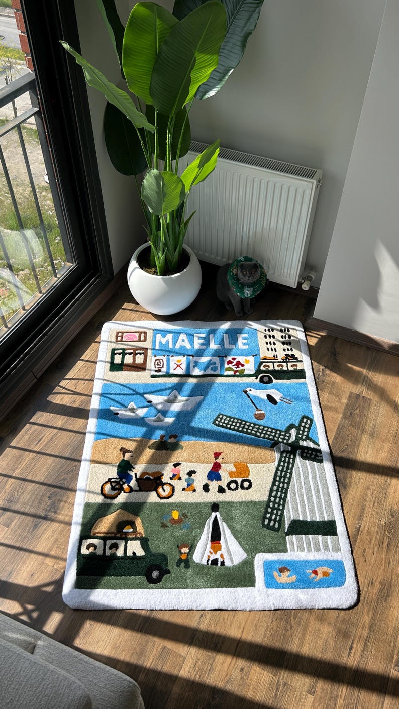 May include: A rectangular rug with a whimsical design featuring a town scene with a windmill, airplane, and people. The rug has a white border and the name "MAELLE" is displayed at the top. The color palette includes blue, green, beige, and brown.