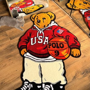 May include: A decorative rug in the shape of a bear wearing a red hoodie with "USA POLO 9" on it, holding a basketball. The bear is wearing cream-colored pants and black and white sneakers. The rug is on a wooden floor.