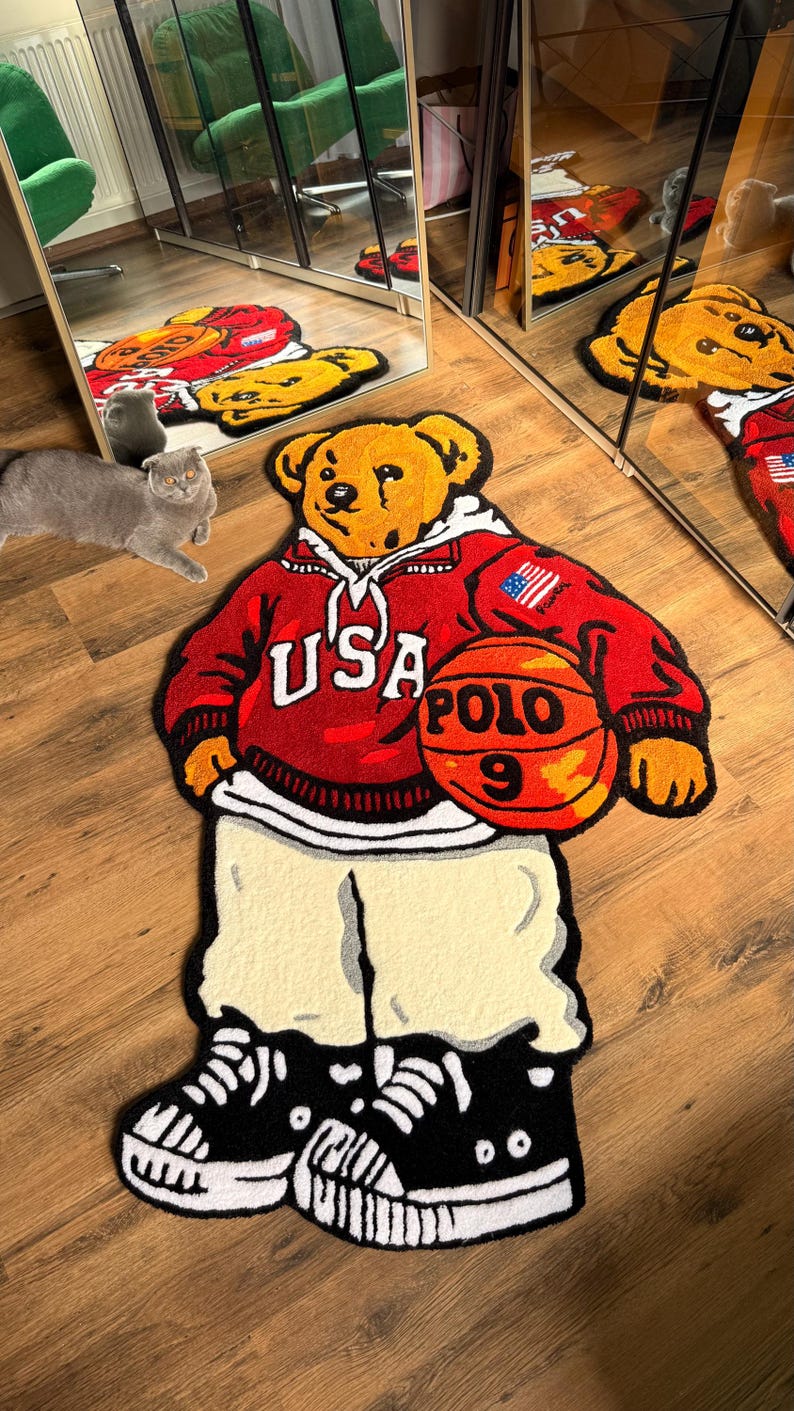May include: A decorative rug in the shape of a bear wearing a red hoodie with "USA POLO 9" on it, holding a basketball. The bear is wearing cream-colored pants and black and white sneakers. The rug is on a wooden floor.