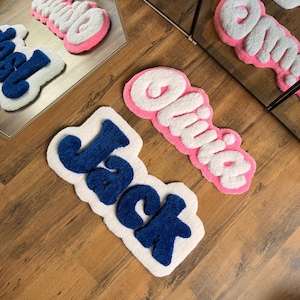 May include: Three custom name rugs. One rug spells "Jack" in blue letters on a white background. Another rug spells "Olivia" in white letters with a pink border. The third rug spells "Olivia" in white letters with a pink border.