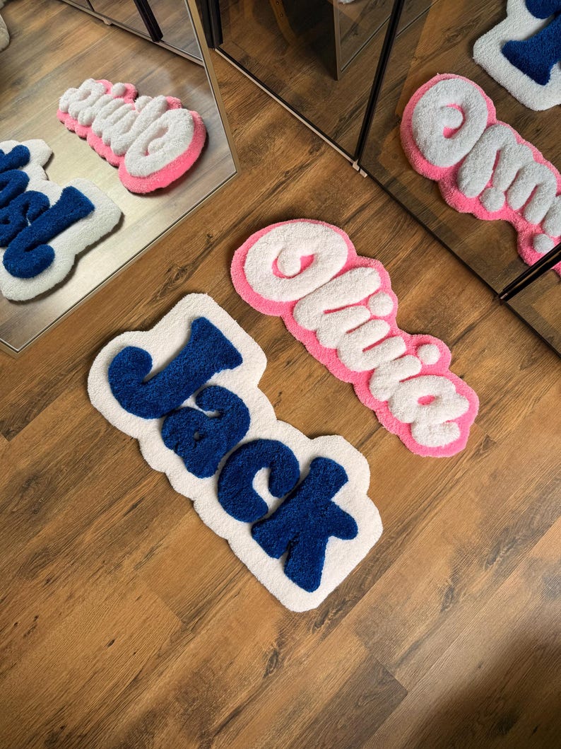 May include: Three custom name rugs. One rug spells "Jack" in blue letters on a white background. Another rug spells "Olivia" in white letters with a pink border. The third rug spells "Olivia" in white letters with a pink border.