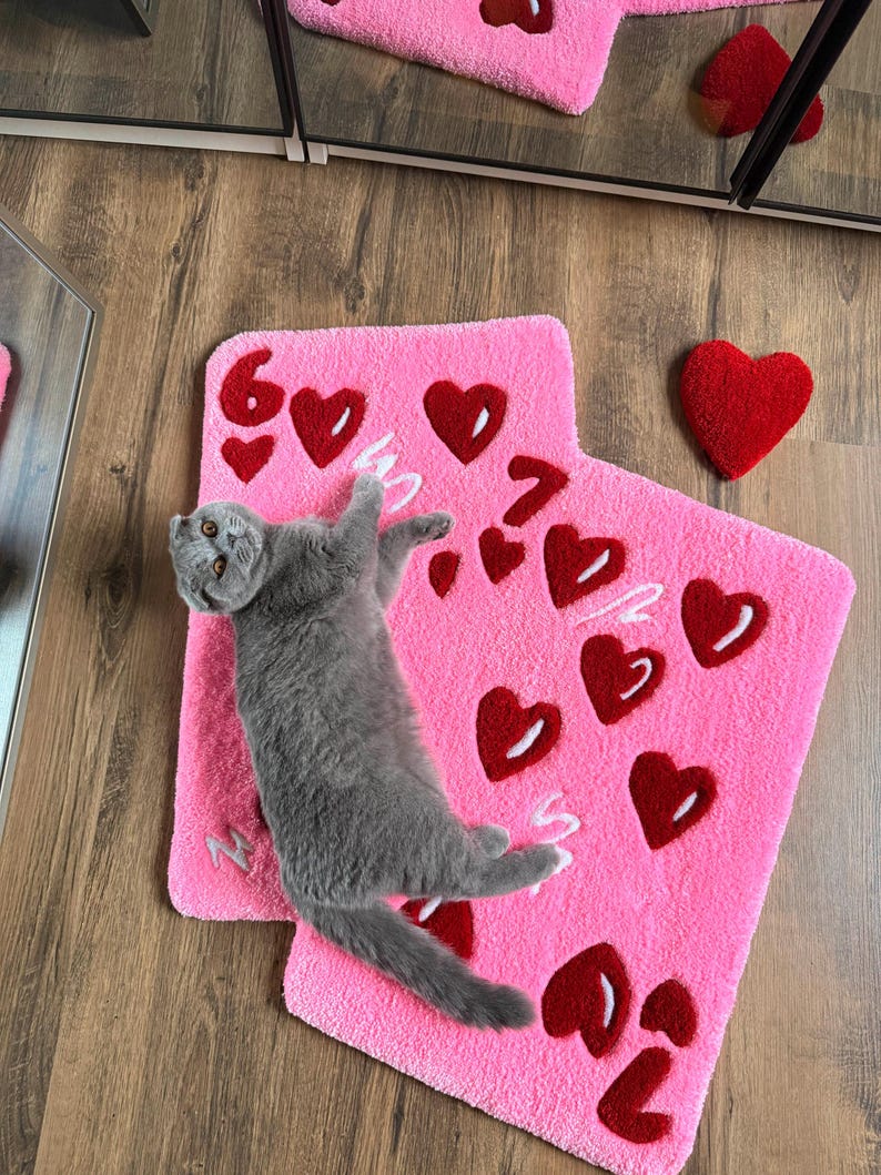 May include: A pink rug with red heart designs and the numbers 6 and 7. A gray cat is lying on the rug. A red heart-shaped pillow is next to the rug. The rug is on a wooden floor.