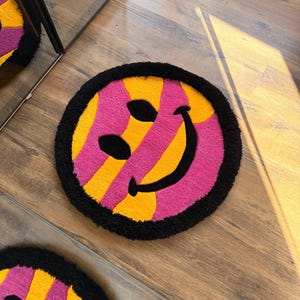 May include: Round rug with a smiley face design. The rug features a black border, a pink and yellow striped pattern, and black eyes and a smile. The rug is on a wooden floor.