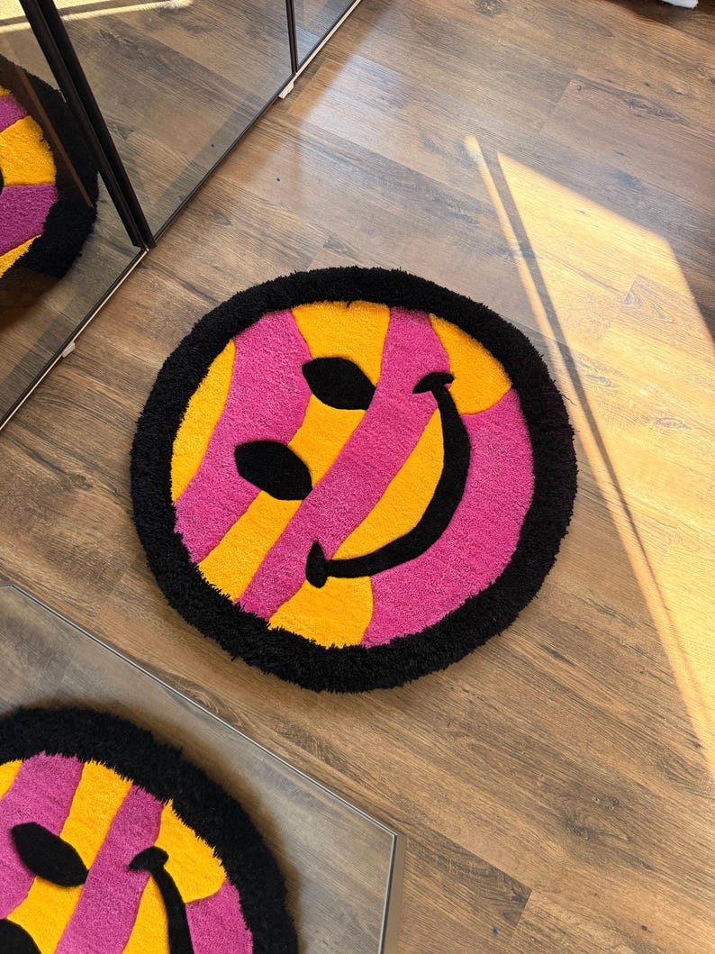 May include: Round rug with a smiley face design. The rug features a black border, a pink and yellow striped pattern, and black eyes and a smile. The rug is on a wooden floor.