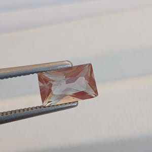May include: A rectangular gemstone, displaying a peach and amber colour gradient, is held by silver tweezers. The gem has a faceted cut, revealing its multiple planes and internal reflections. The background is a soft, neutral white.