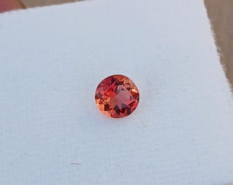 May include: A round, faceted gemstone with a vibrant orange-red hue. The gem is set against a white background, showcasing its color and clarity. The gemstone's cut allows light to reflect, creating a sparkling effect.