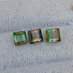 May include: Three square-cut gemstones featuring a gradient of green, yellow, and pink hues. Each stone displays a faceted surface with translucent clarity, arranged in a horizontal row, showcasing a distinct color transition within each individual gem.