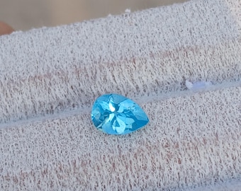 May include: A pear-shaped, faceted gemstone in a vibrant, light blue hue. The gem is displayed on a textured, off-white fabric, showcasing its clarity and brilliance. The gemstone's cut allows light to reflect, creating a sparkling effect.