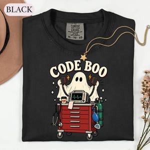 May include: A black t-shirt featuring a graphic of a ghost wearing a stethoscope and holding defibrillator paddles above a red medical cart. The text CODE BOO is printed in cream above the ghost. The cart includes a monitor, oxygen tank, and coffee cup.