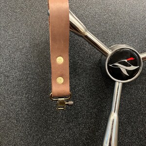 May include: A brown leather strap with brass rivets and a metal clasp is attached to a chrome handle with a black face. The black face has a white bird graphic with a red accent.