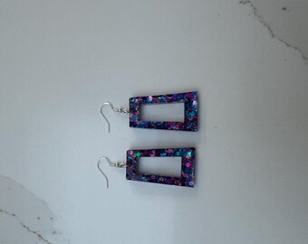 May include: A pair of rectangular dangle earrings. The earrings are dark blue with a glittery, colourful interior. The earrings have a rectangular cutout in the centre and silver-coloured ear wires.
